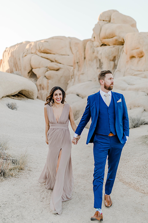 bride in a taupe maxi gown with straps and her hair down, the groom in a cobalt blue suit with a neutral long tie, walking on dune