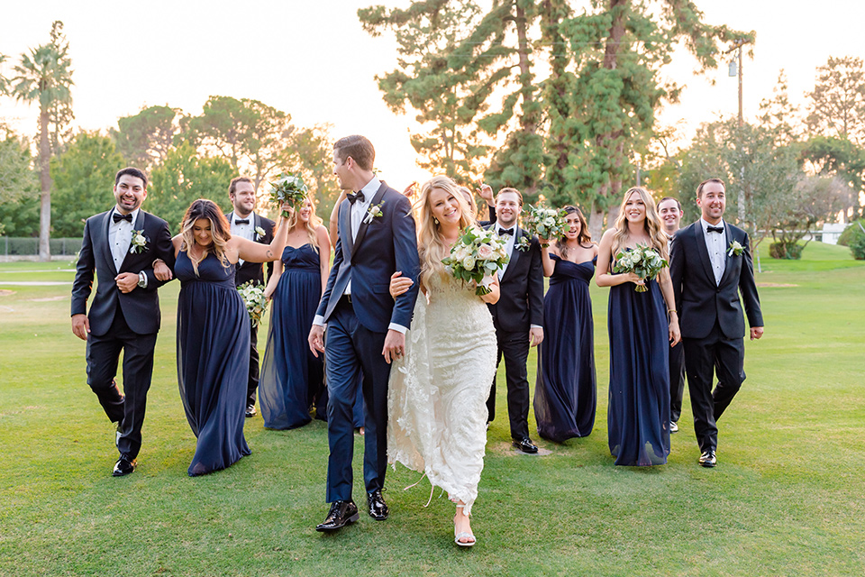  bride in a formfitting gown with thin straps and lace details, the groom in a navy tuxedo, the bridesmaids in black off-the-shoulder gowns, and the groomsmen in black tuxedos 