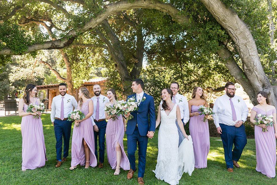  bride in a lace form-fitting gown with straps, the groom in a dark blue suit with a white long tie, the bridesmaids in lavender gowns, the groomsmen in blue pants and lavender long ties
