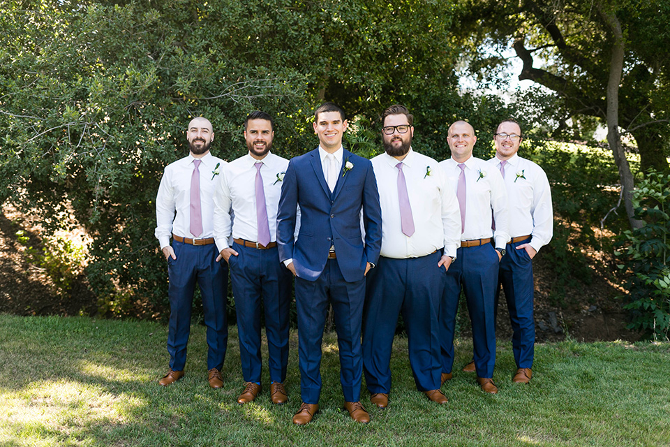  the groom in a dark blue suit with a white long tie and the groomsmen in blue pants and lavender long ties