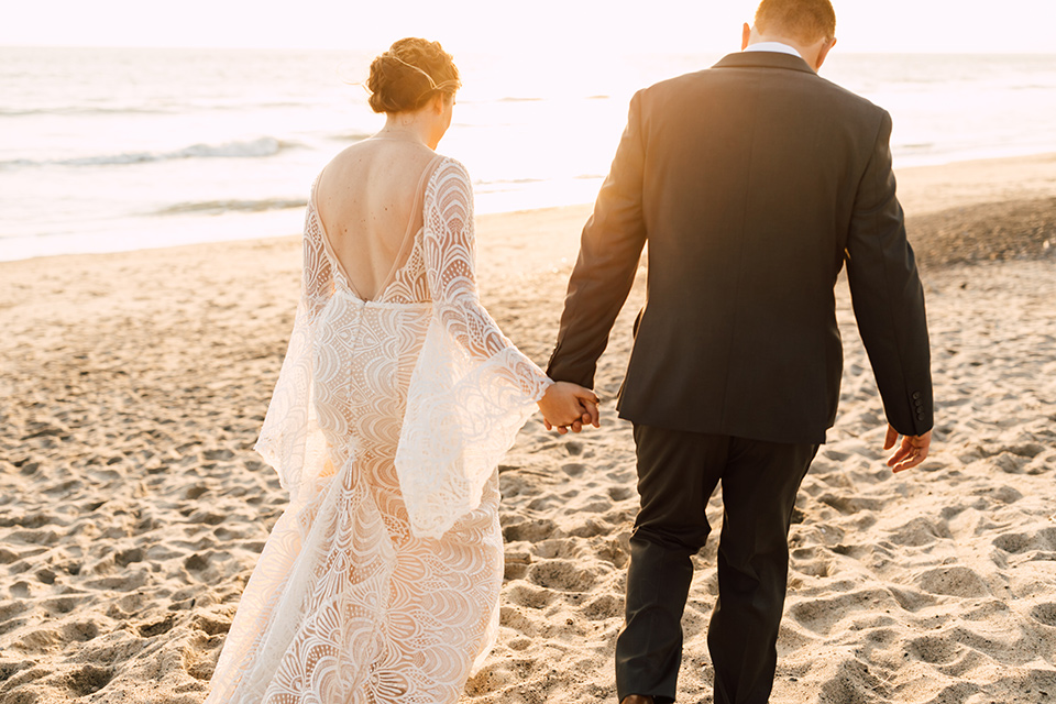bride in a lace bohemian gown with a high neckline and flutter sleeves, the groom was in grey retail suit with a blue long tie, walking on sand