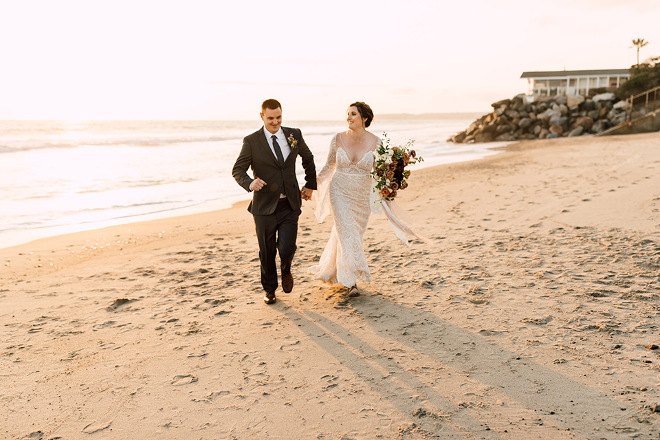 bride in a lace bohemian gown with a high neckline and flutter sleeves, the groom was in grey retail suit with a blue long tie, walking on sand