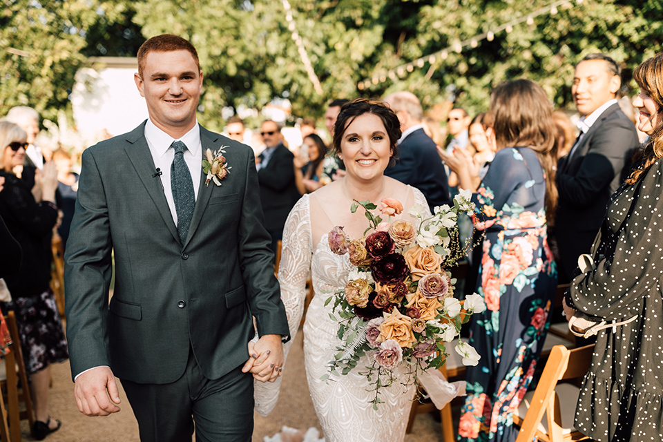 bride in a lace bohemian gown with a high neckline and flutter sleeves, the groom was in grey retail suit with a blue long tie, walking down the aisle