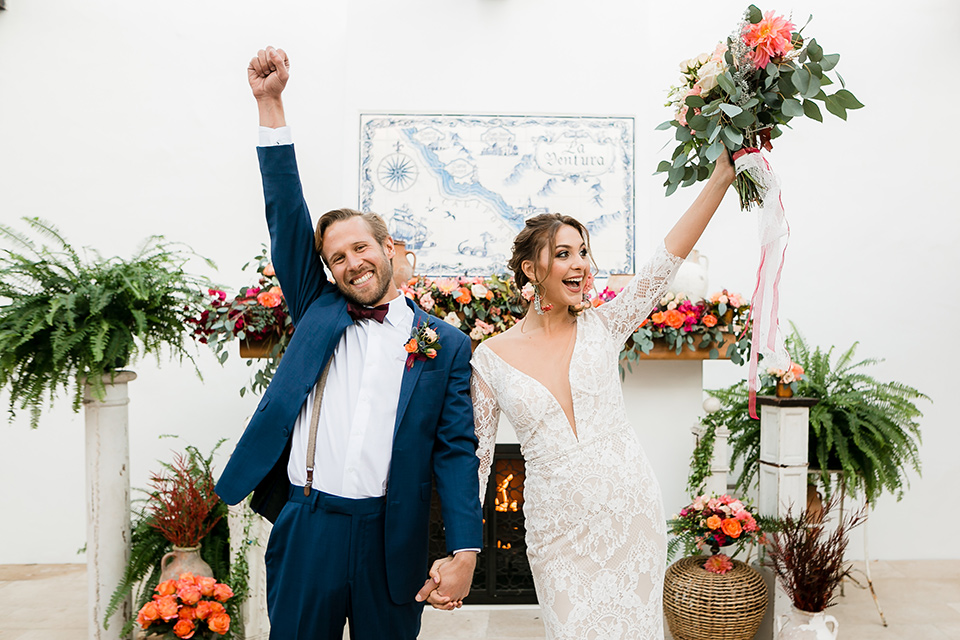  bride in a Spanish inspired lace formfitting gown with and the groom in a dark blue suit with a burgundy bow tie and suspenders, cheering after the ceremony