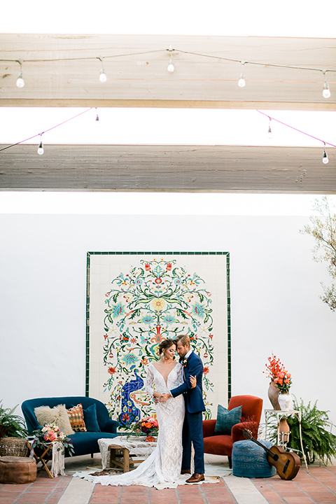  Spanish inspired venue with blue furniture and the bride and groom sitting in the venue, the bride in a lace formfitting gown with sleeves and dark blue suit