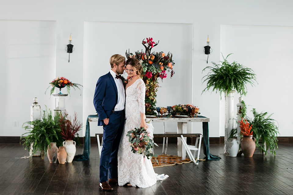  bride in a Spanish inspired lace formfitting gown with and the groom in a dark blue suit with a burgundy bow tie and suspenders, standing in the venue