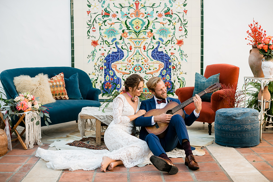  bride in a Spanish inspired lace formfitting gown with and the groom in a dark blue suit with a burgundy bow tie and suspenders, sitting in the venue with the guitar