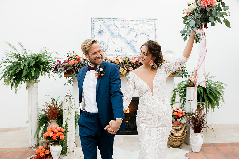  bride in a Spanish inspired lace formfitting gown with and the groom in a dark blue suit with a burgundy bow tie and suspenders, walking down the aisle