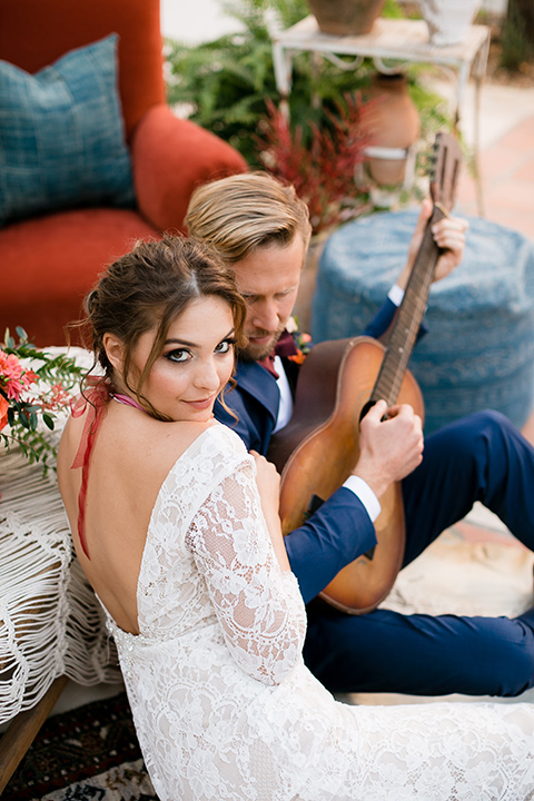  the bride in a lace formfitting gown with sleeves and a floral necklace, the groom in a dark blue suit with a burgundy bow tie sitting and playing a guitar