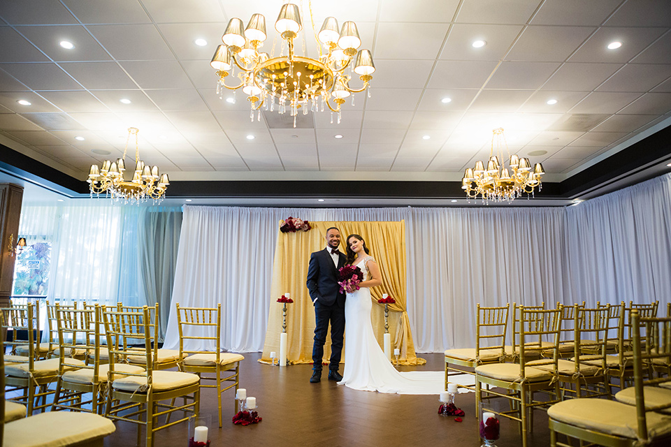  bride in a satin white formfitting gown with a plunging neckline and a trumpet train skirt holding a bouquet of purple flowers the groom in a navy tuxedo with a black satin lapel and a purple bow tie at the ceremony