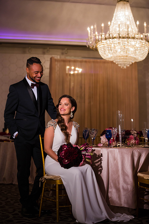 bride with a soft finger wave hair style with delicate makeup and gold earrings, the groom in a navy tuxedo with a black satin lapel and a purple bow tie sitting at the table