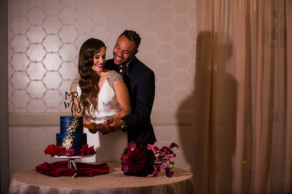  bride in a satin white formfitting gown with a plunging neckline and a trumpet train skirt holding a bouquet of purple flowers the groom in a navy tuxedo with a black satin lapel and a purple bow tie at cutting the cake