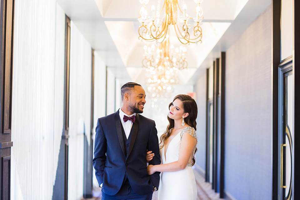  bride in a satin white formfitting gown with a plunging neckline and a trumpet train skirt holding a bouquet of purple flowers the groom in a navy tuxedo with a black satin lapel and a purple bow tie smiling at each other in the hallway