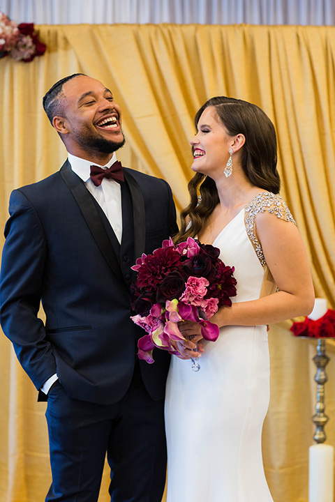 bride with a soft finger wave hair style with delicate makeup and gold earrings, the groom in a navy tuxedo with a black satin lapel and a purple bow tie laughing