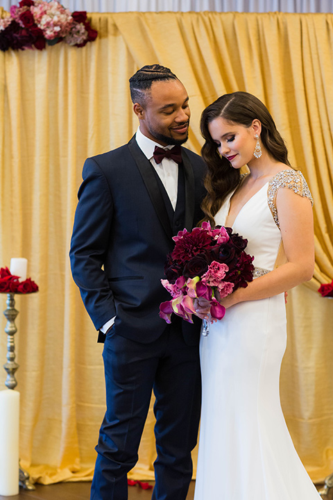  bride with a soft finger wave hair style with delicate makeup and gold earrings, the groom in a navy tuxedo with a black satin lapel and a purple bow tie smiling at each other 