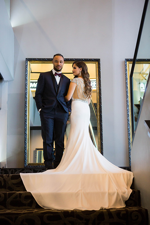 bride with a soft finger wave hair style with delicate makeup and gold earrings, the groom in a navy tuxedo with a black satin lapel and a purple bow tie at the top of the stairs