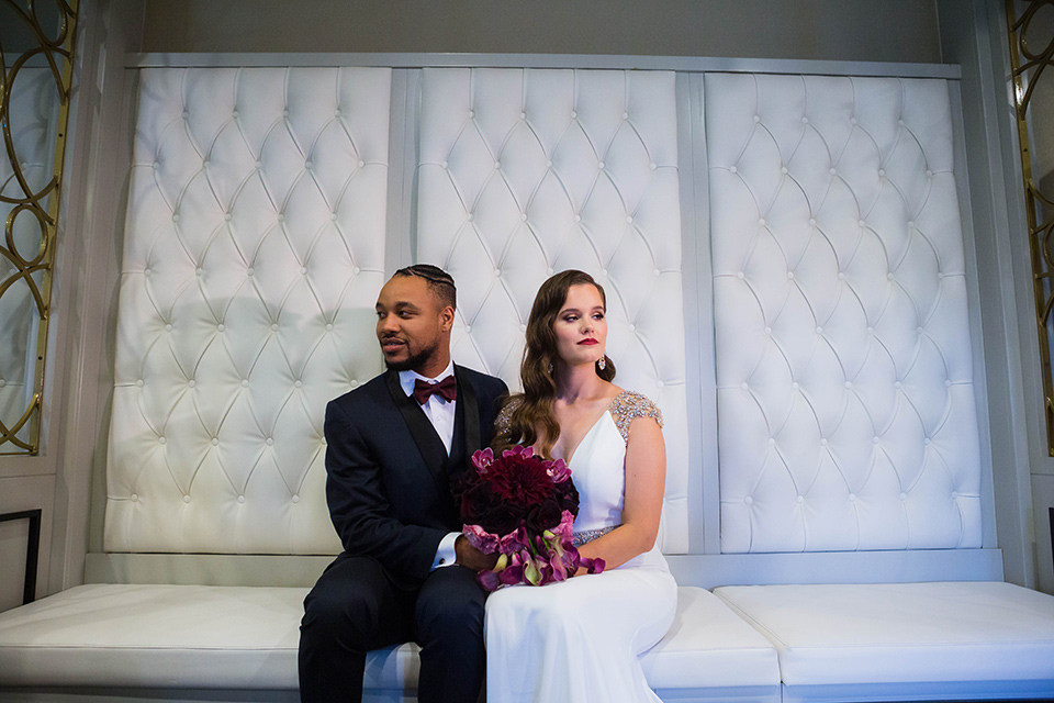  bride in a satin white formfitting gown with a plunging neckline and a trumpet train skirt holding a bouquet of purple flowers the groom in a navy tuxedo with a black satin lapel and a purple bow tie sitting on the white regal couch