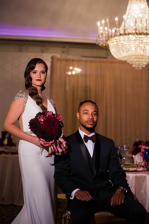  bride with a soft finger wave hair style with delicate makeup and gold earrings, the groom in a navy tuxedo with a black satin lapel and a purple bow tie sitting at the table 