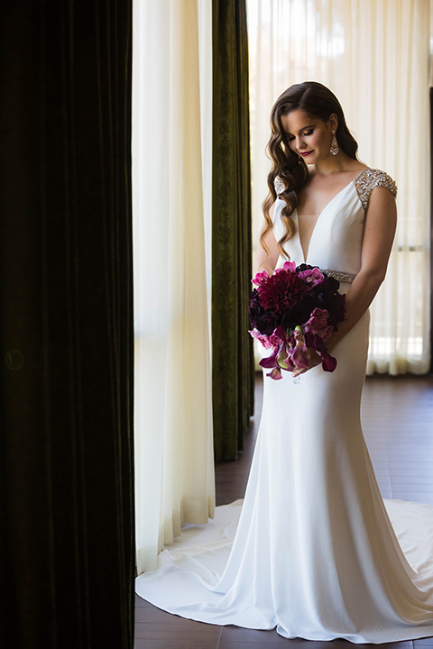  bride in a satin white formfitting gown with a plunging neckline and a trumpet train skirt holding a bouquet of purple flowers