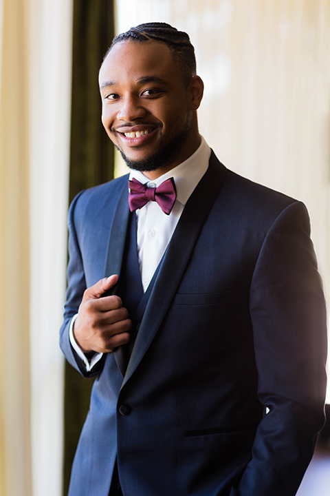  the groom in a navy tuxedo with a black satin lapel and a purple bow tie holding the lapel of his jacket