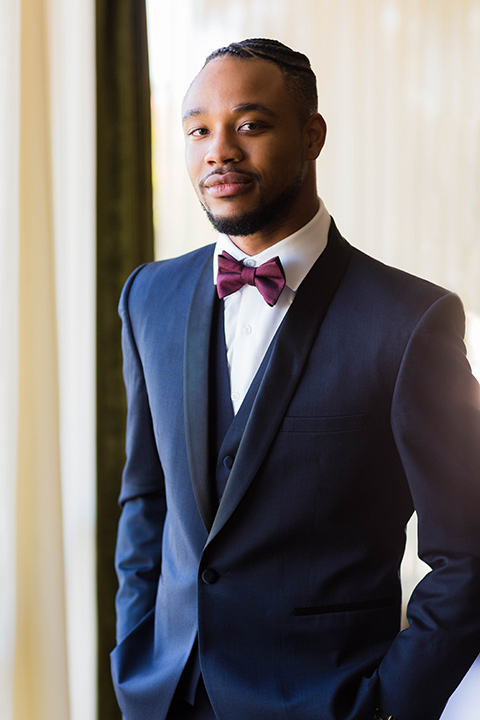  the groom in a navy tuxedo with a black satin lapel and a purple bow tie smiling at camera 