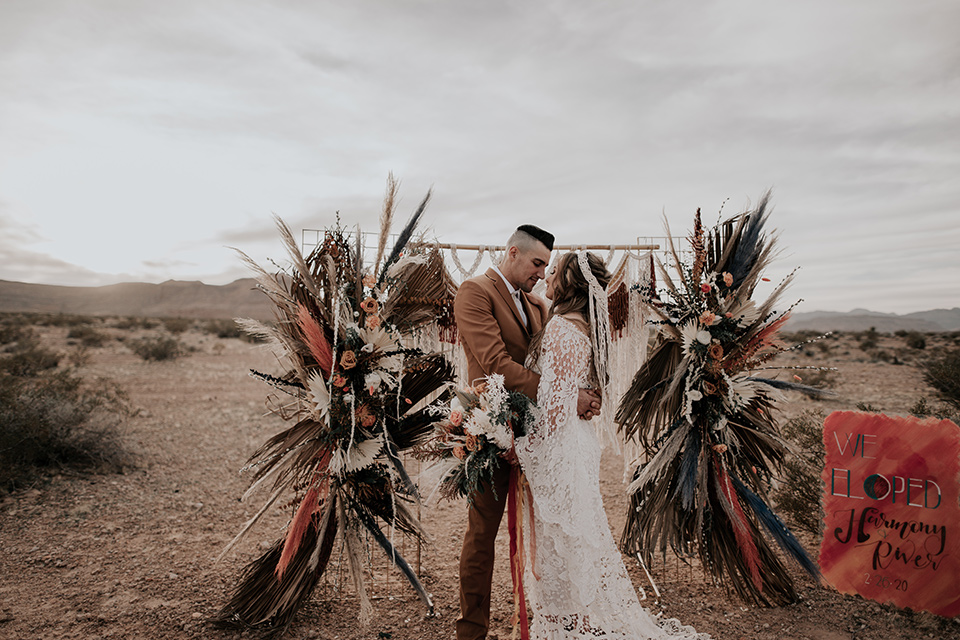  bride in a bohemian gown with long sleeves and lace detailing and the groom in a caramel suit with a brown long tie and a fun colored ceremony arch with bright colored pampas grass 