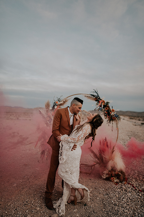  bride in a bohemian white gown with lace detailing and long sleeves and the groom in a caramel rust suit color with a brown long tie, they are dancing in with pink smoke around them