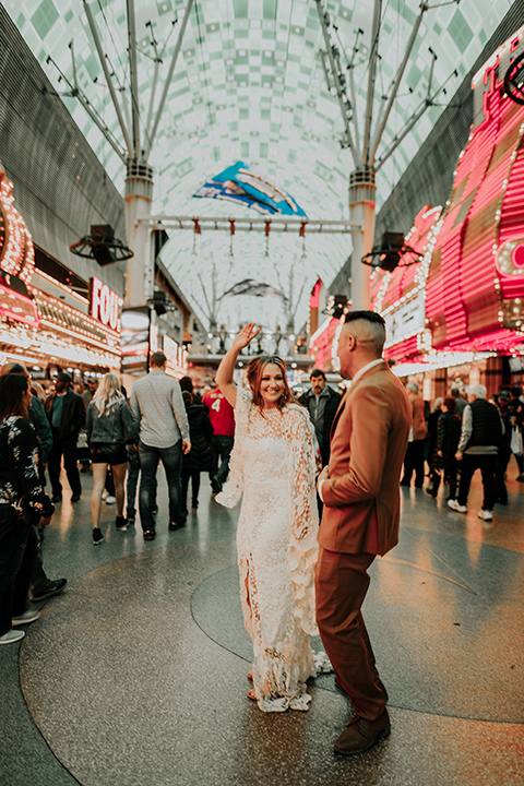  bride in a bohemian white gown with lace detailing and long sleeves and the groom in a caramel rust suit color with a brown long tie, smiling downtown 