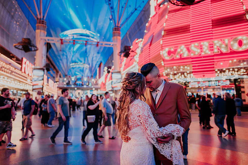  bride in a bohemian gown with long sleeves and lace detailing and the groom in a caramel suit with a brown long tie kissing downtown 