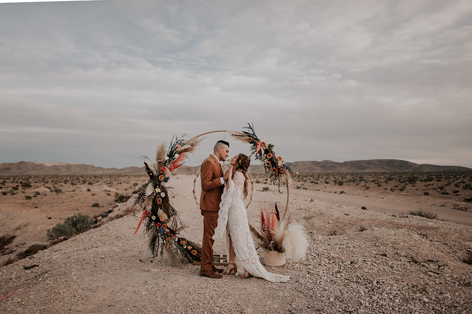  bride in a bohemian gown with long sleeves and lace detailing and the groom in a caramel suit with a brown long tie at ceremony 
