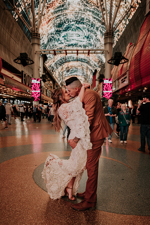  bride in a bohemian white gown with lace detailing and long sleeves and the groom in a caramel rust suit color with a brown long tie, kissing downtown 