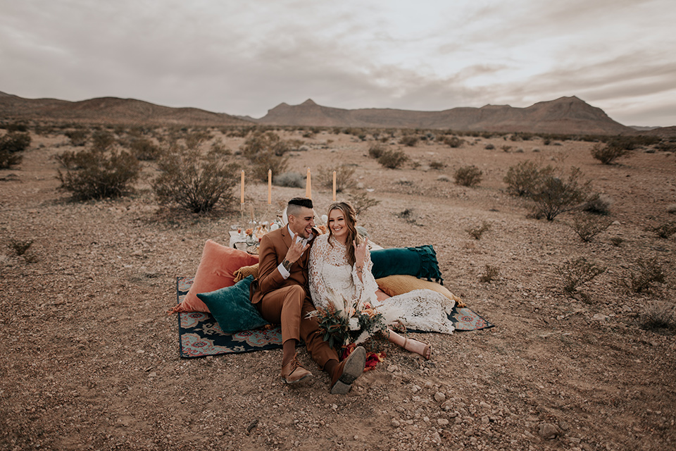  bride in a bohemian gown with long sleeves and lace detailing and the groom in a caramel suit with a brown long tie sitting on picnic tables on the ground 