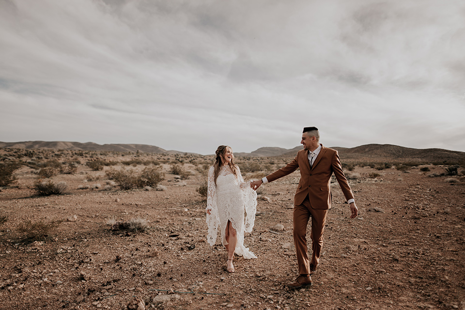  bride in a bohemian gown with long sleeves and lace detailing and the groom in a caramel suit with a brown long tie walking in the desert 