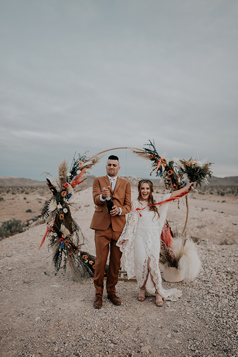  bride in a bohemian white gown with lace detailing and long sleeves and the groom in a caramel rust suit color with a brown long tie, opening champagne