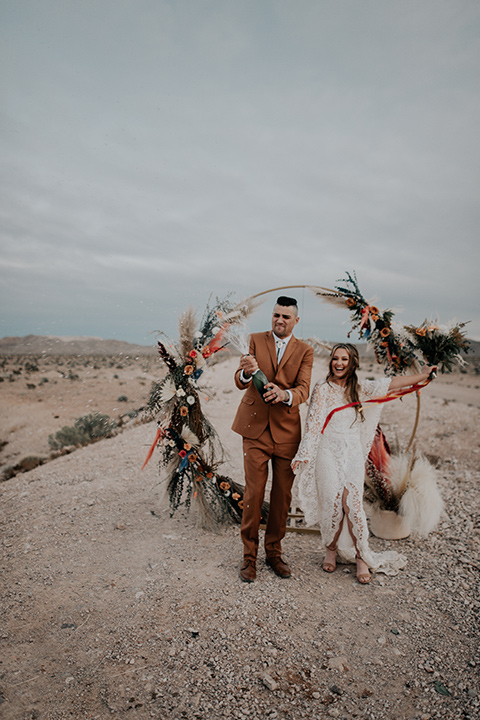  bride in a bohemian white gown with lace detailing and long sleeves and the groom in a caramel rust suit color with a brown long tie, opening champagne 