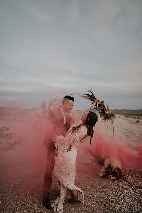  bride in a bohemian white gown with lace detailing and long sleeves and the groom in a caramel rust suit color with a brown long tie, they are dancing in with pink smoke around them 