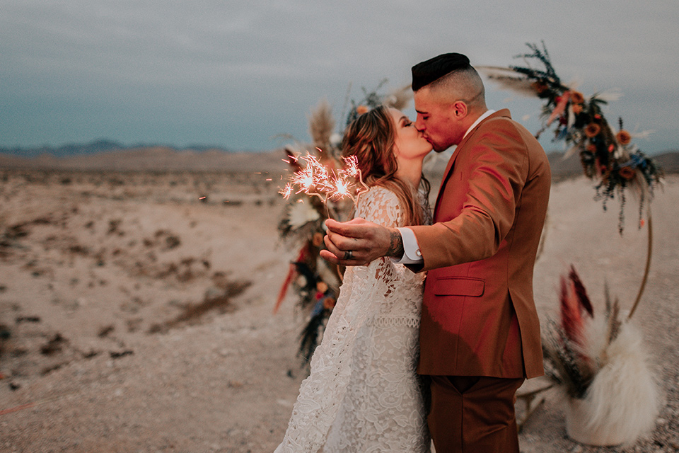  bride in a bohemian gown with long sleeves and lace detailing and the groom in a caramel suit with a brown long tie with sparklers kissing 