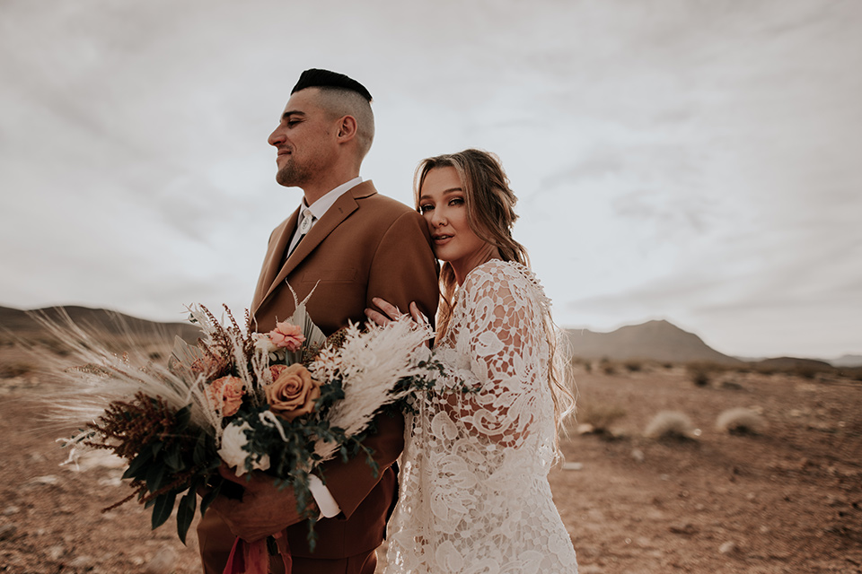  bride in a bohemian gown with long sleeves and lace detailing and the groom in a caramel suit with a brown long tie hugging 