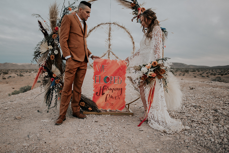  bride in a bohemian gown with long sleeves and lace detailing and the groom in a caramel suit with a brown long tie at ceremony 