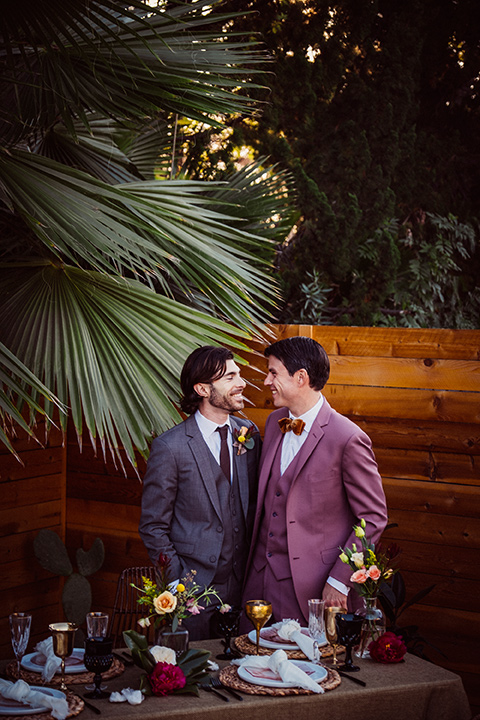  groom in a café brown suit and a brown long tie and the other groom in a rose pink suit with a gold velvet bow tie, outside at the reception 