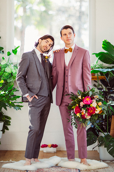  groom in a café brown suit and a brown long tie and the other groom in a rose pink suit with a gold velvet bow tie, outside at the ceremony 