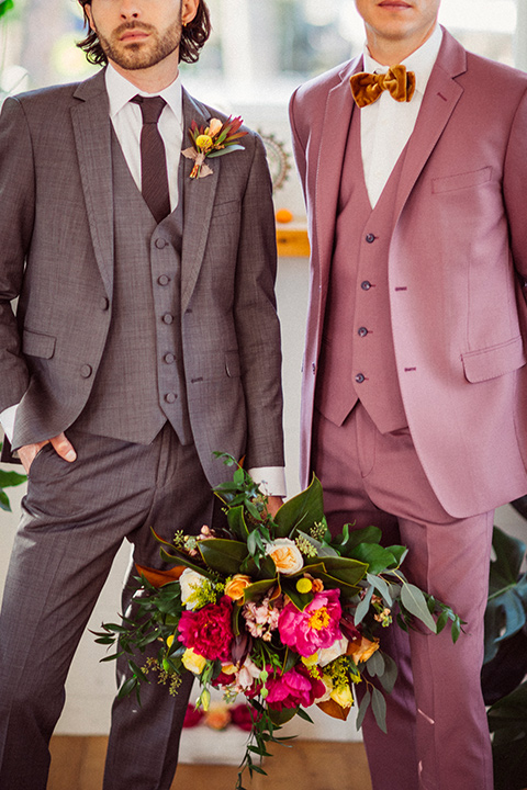  groom in a café brown suit and a brown long tie and the other groom in a rose pink suit with a gold velvet bow tie, outside at the ceremony 