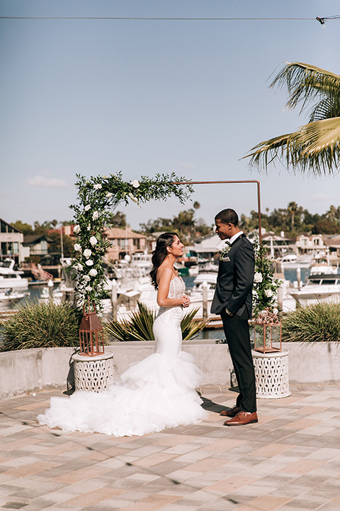  bride in a lace white gown with a long veil and the groom in a navy shawl lapel tuxedo with black bow ties, and the bridesmaid in a silver long gown and the groomsmen in a navy shawl lapel tuxedo with a black bow tie on the beach