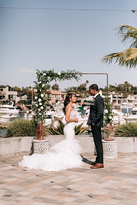  bride in a lace white gown with a long veil and the groom in a navy shawl lapel tuxedo with black bow ties, and the bridesmaid in a silver long gown and the groomsmen in a navy shawl lapel tuxedo with a black bow tie on the beach