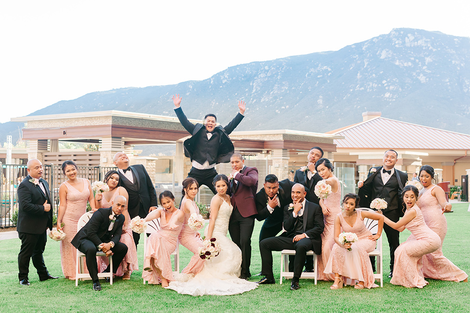  bride in a lace formfitting gown and a high neckline and the groom in a burgundy tuxedo with a black bow tie, and the bridesmaids in a rose blush gowns and the groomsmen in a black tuxedo with a black bow tie 