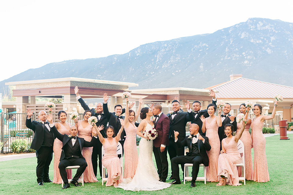  bride in a lace formfitting gown and a high neckline and the groom in a burgundy tuxedo with a black bow tie, and the bridesmaids in a rose blush gowns and the groomsmen in a black tuxedo with a black bow tie 