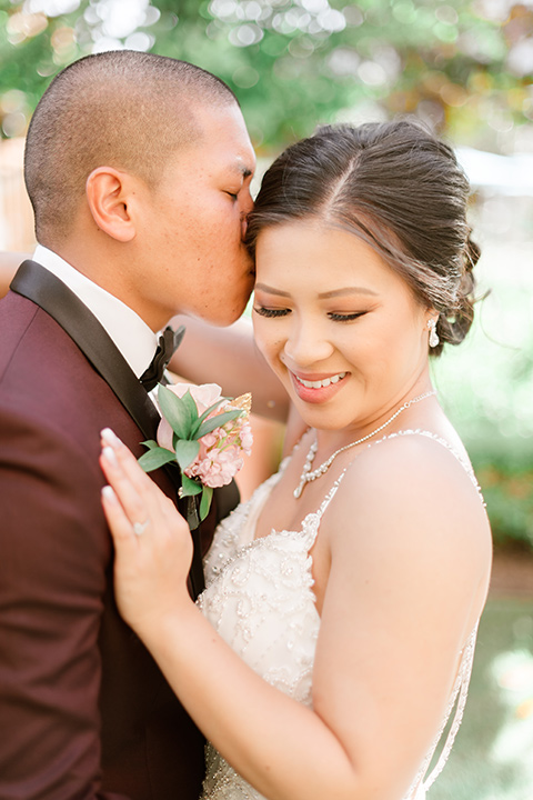  bride in a lace formfitting gown and a high neckline and the groom in a burgundy tuxedo with a black bow tie close together