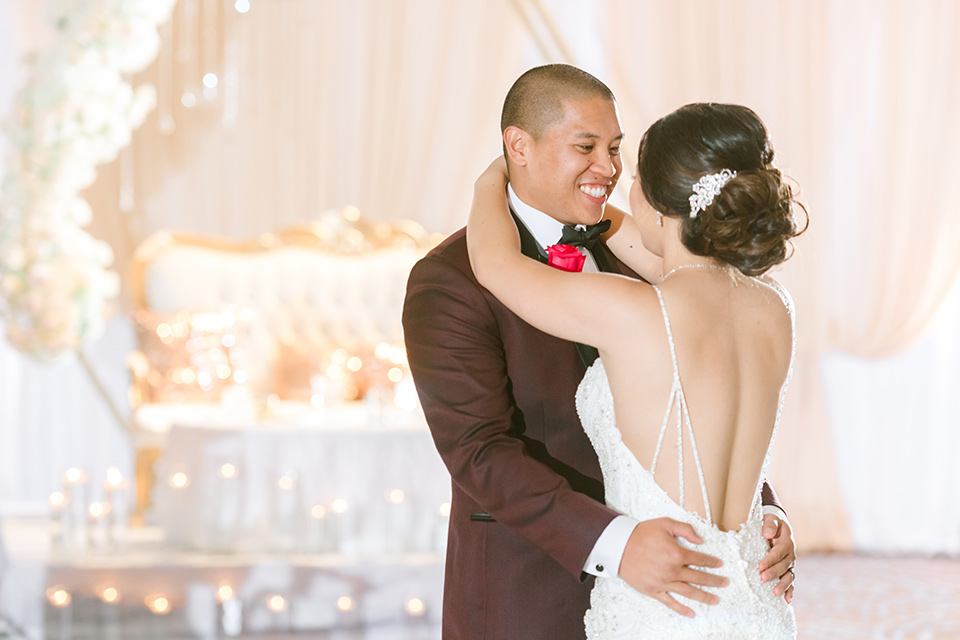  bride in a lace formfitting gown and a high neckline and the groom in a burgundy tuxedo with a black bow tie dancing at the reception