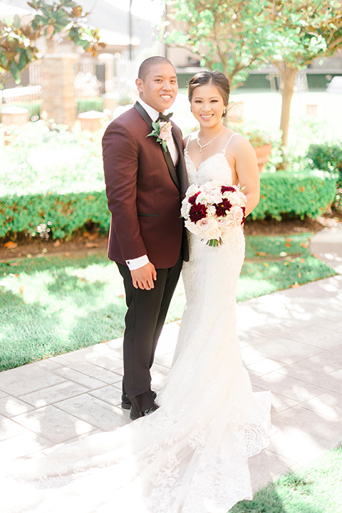  bride in a lace formfitting gown and a high neckline and the groom in a burgundy tuxedo with a black bow tie smiling 