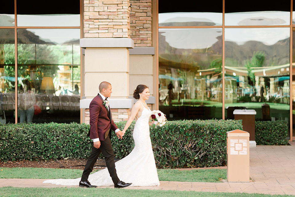  bride in a lace formfitting gown and a high neckline and the groom in a burgundy tuxedo with a black bow tie walking